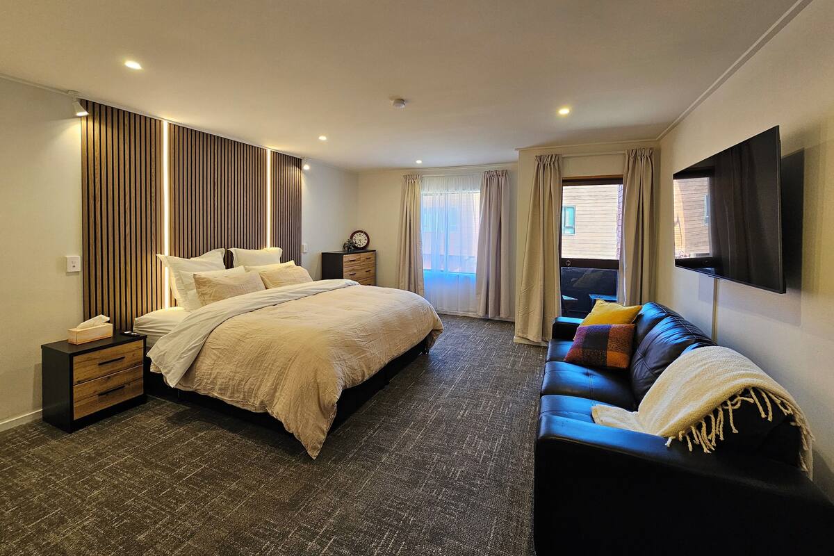 A modern hotel room with a large bed, a sofa, a TV, and a window with curtains.