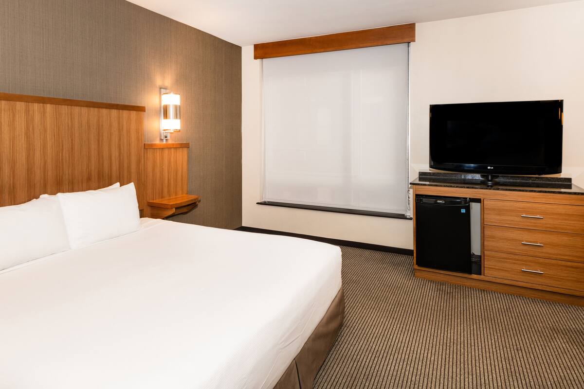 Ein Hotelzimmer mit einem großen Bett, einem Fernseher, einem kleinen Nachttisch und einem Fenster mit Jalousien.