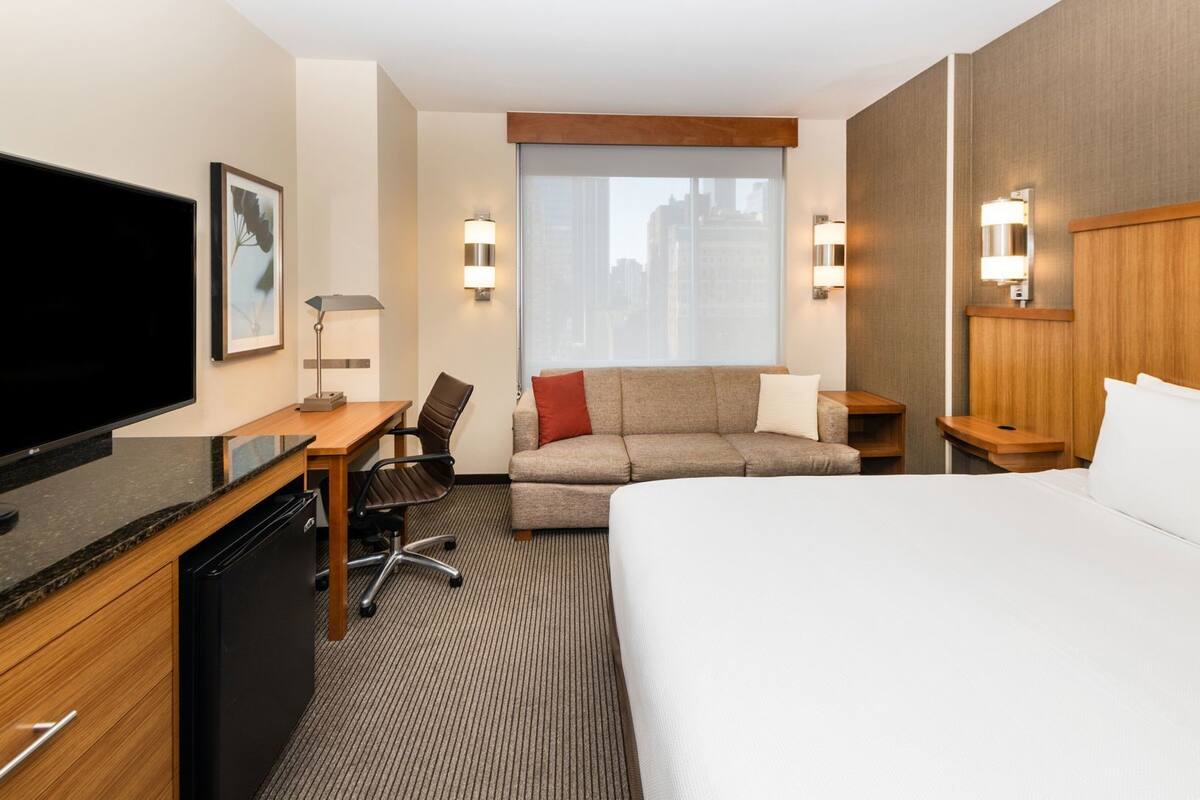 Une chambre d’hôtel comprenant un lit, un bureau avec un ordinateur, un canapé, une télévision et une fenêtre donnant sur la ville.