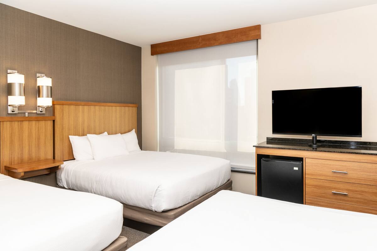 Ein Hotelzimmer mit zwei Betten, einem Fernseher, einem Kühlschrank und einem Fenster mit Jalousien.