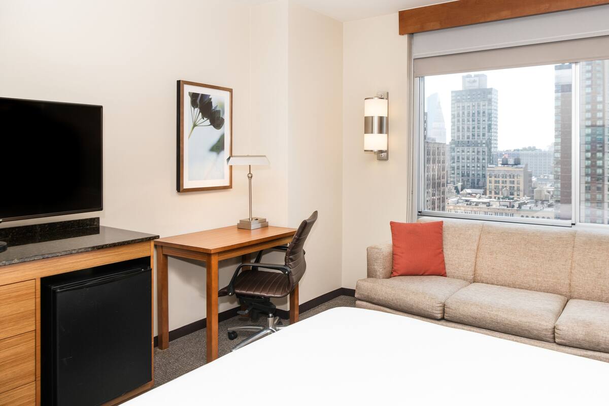 Ein Hotelzimmer mit Bett, Schreibtisch, Stuhl, Fernseher, einem gerahmten Bild, Lampe, Sofa und einem Fenster mit Stadtblick.