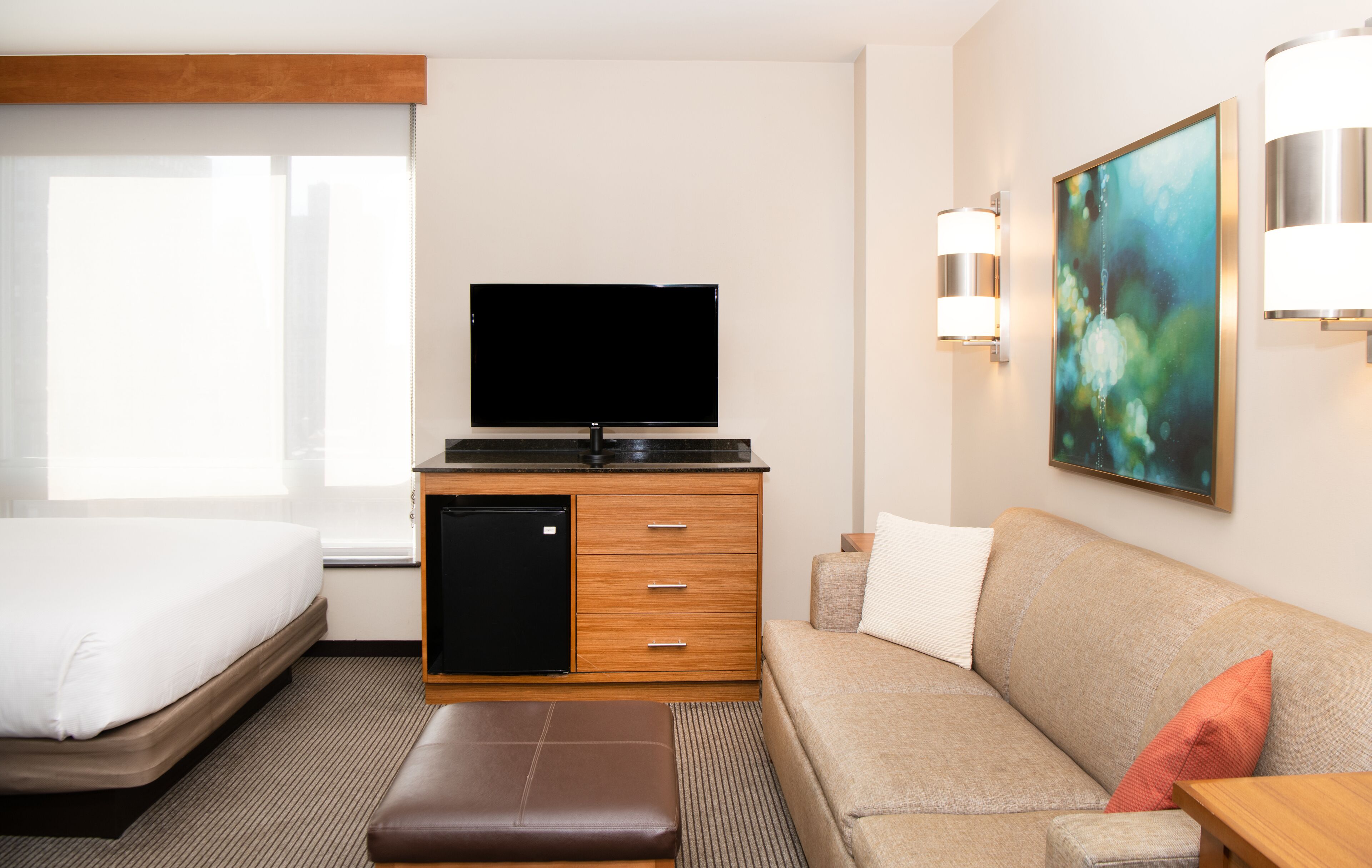 Habitación de hotel con cama, sofá, televisor, refrigerador pequeño y un cuadro en la pared.