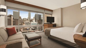 Hotelkamer met groot raam en uitzicht op de stad, een bed, een bank, een televisie en een kleine tafel.