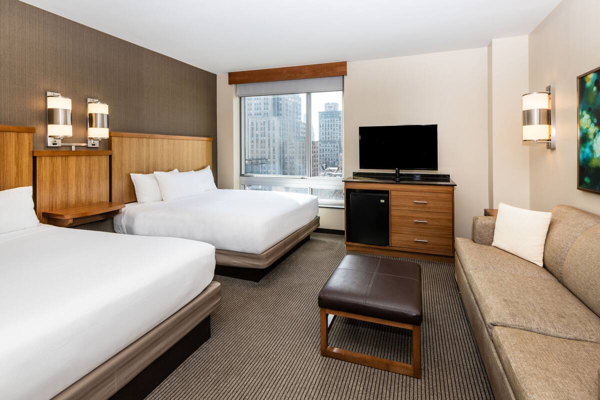 Ein Hotelzimmer mit zwei Betten, einem Fernseher, einem Sofa und einem Fenster mit Blick auf die Stadt.