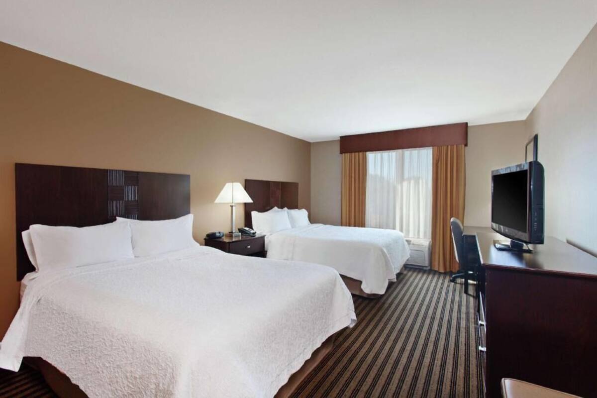 A hotel room with two beds, a desk with a television, a lamp, and a window with curtains.