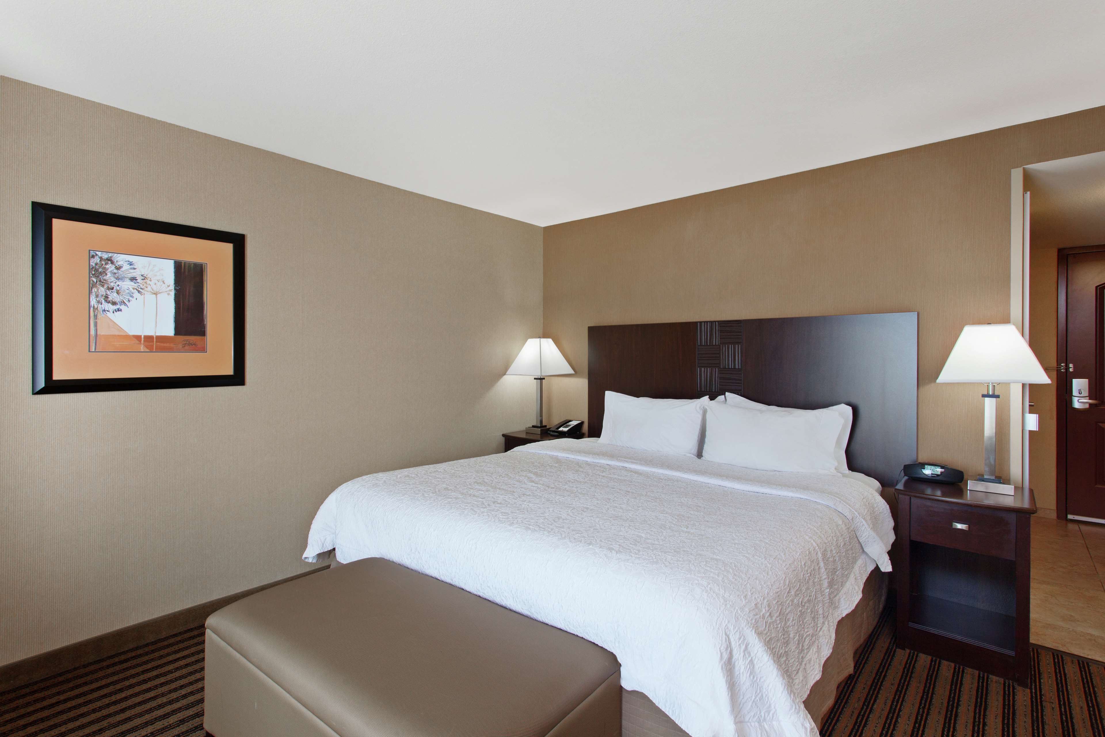 Una habitación de hotel con una cama grande, dos mesitas de noche con lámparas, un cuadro enmarcado en la pared y un banco al pie de la cama.
