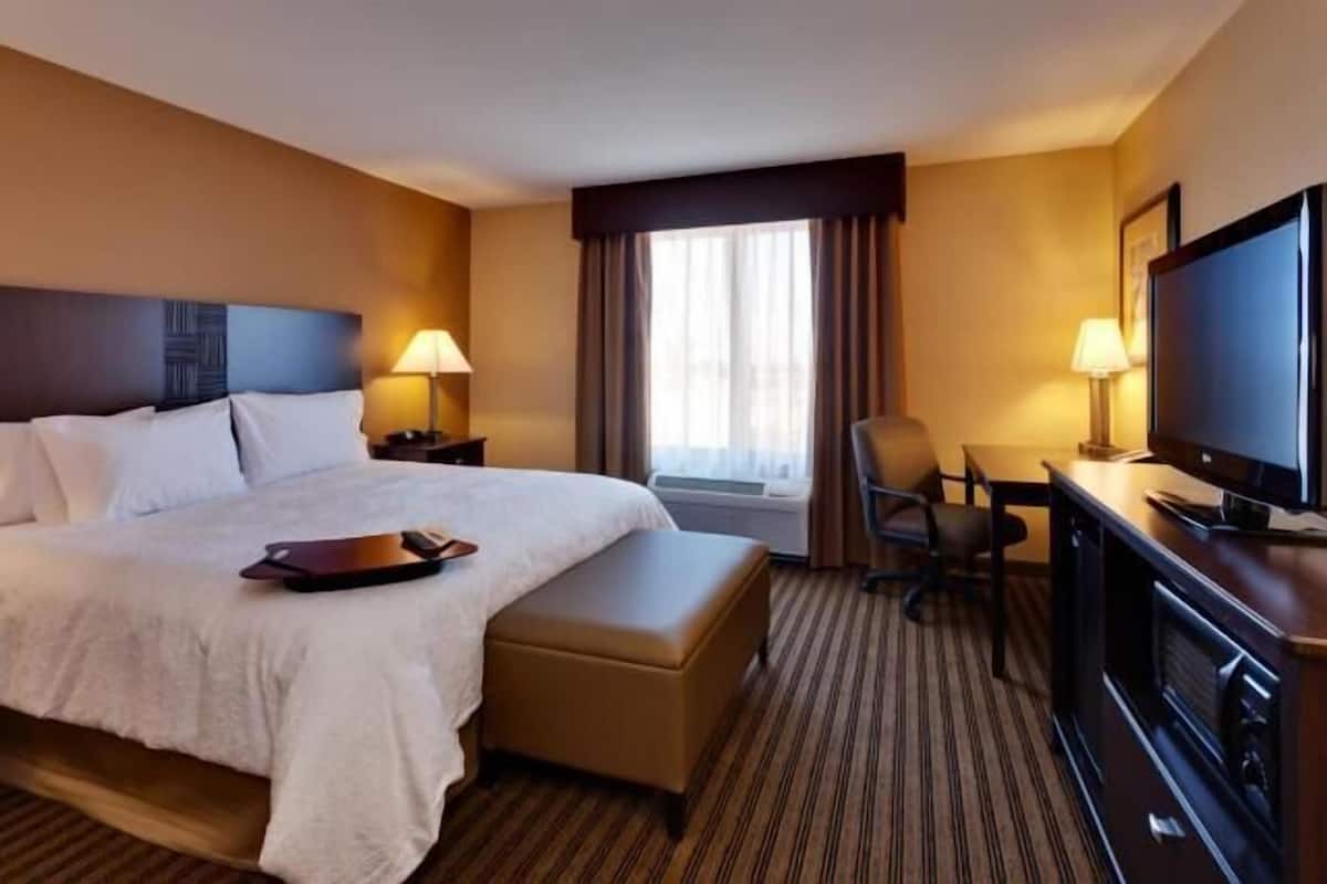 Quarto de hotel com uma cama grande, uma mesa com televisão, uma cadeira e uma janela com cortinas.
