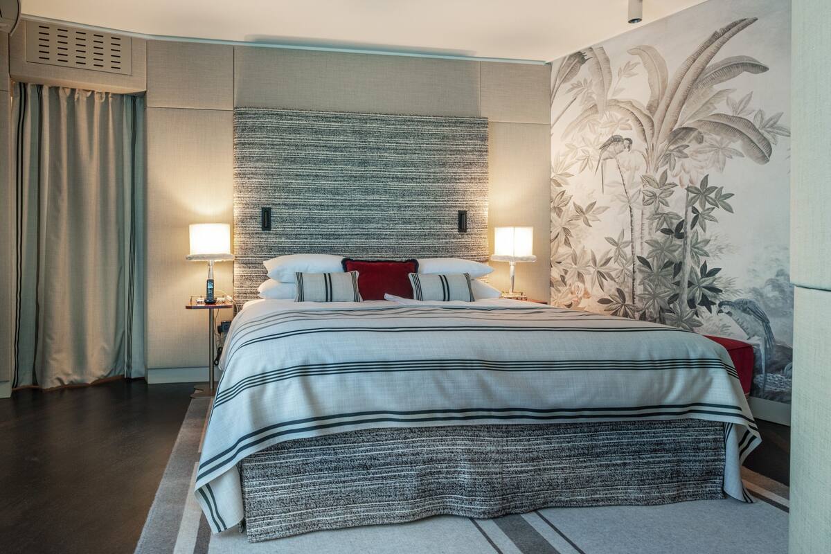 A hotel room with a large bed, two bedside tables, a wall-mounted air conditioning unit, and a large floral mural on the wall.