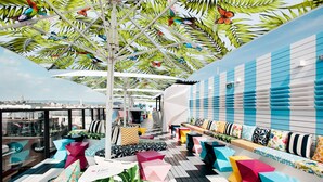 A rooftop bar with colorful seating, a view of the city, and a tropical-themed ceiling.