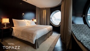 A hotel room with a large bed, two bedside tables with lamps, a round window with a view, and a small sofa.
