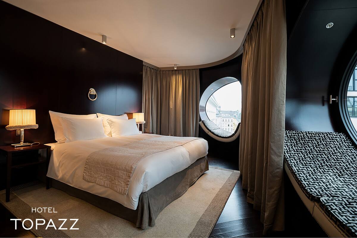 A hotel room with a large bed, two bedside tables with lamps, a round window with a view, and a small sofa.