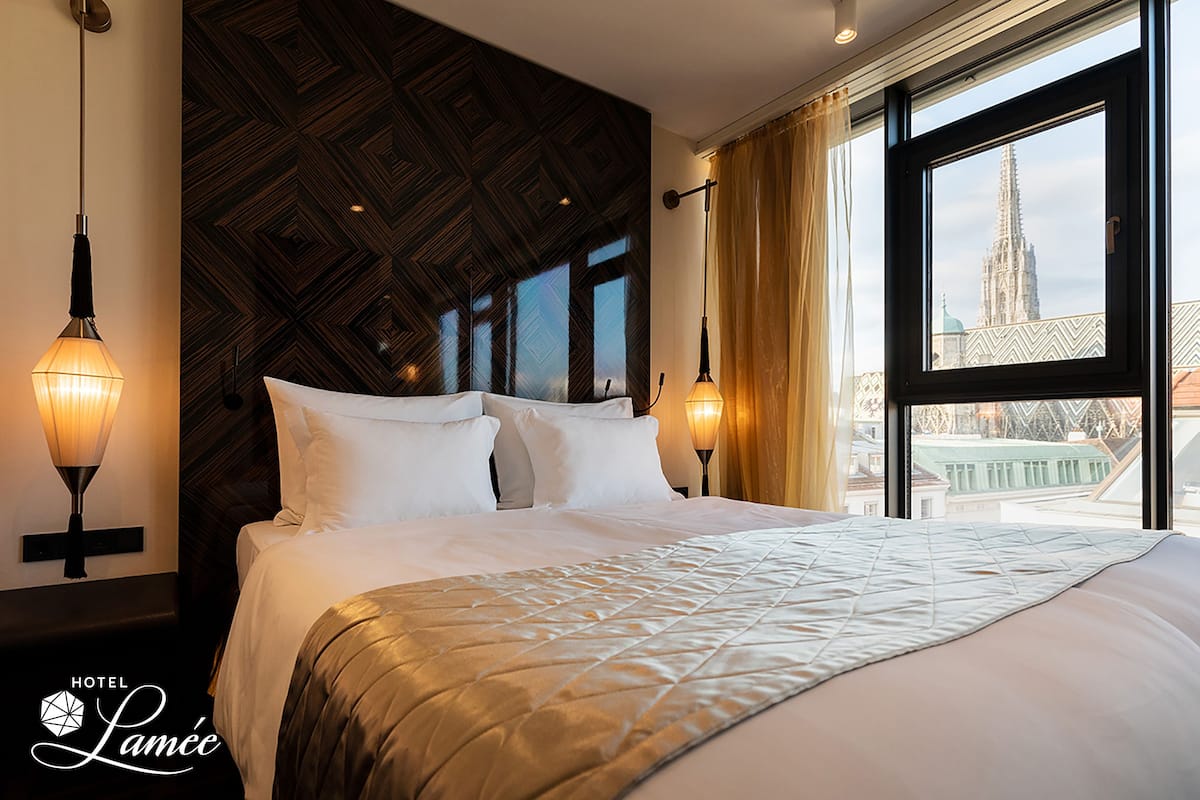 A hotel room with a large bed, two wall-mounted lamps, and a window with a view of a building.