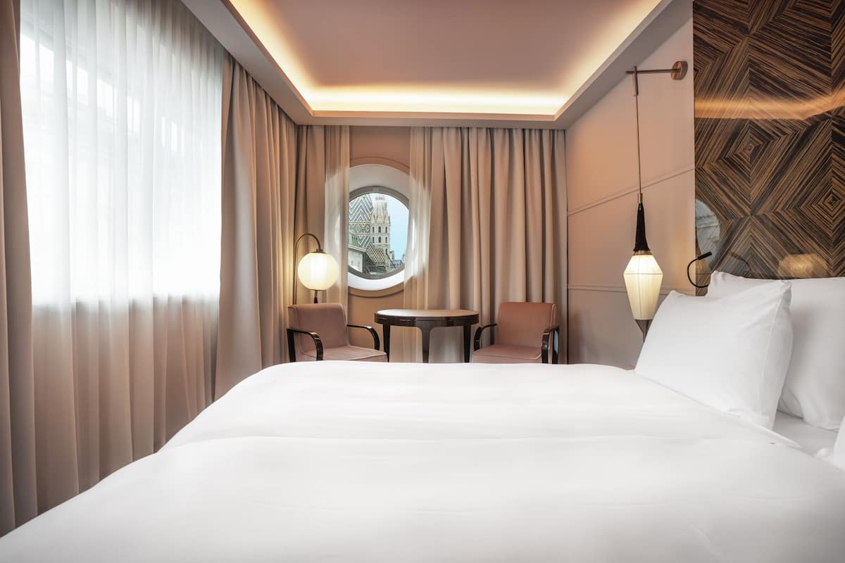A hotel room with a large bed, two chairs, a small table, a round window, and a patterned wall.
