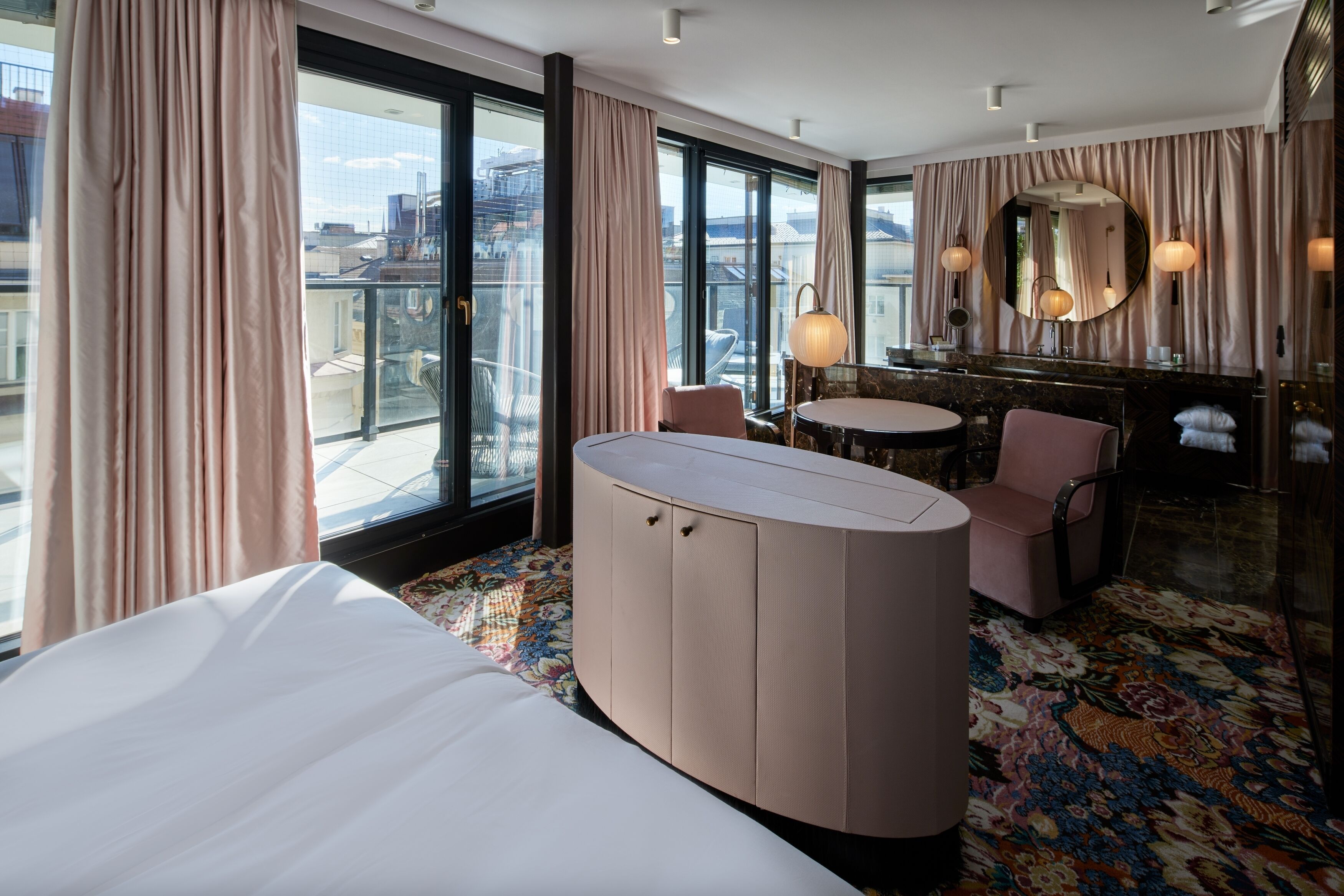 Een hotelkamer met een groot bed, een gebogen kaptafel, een ronde tafel en een stoel. Er is een balkon met uitzicht op de stad.