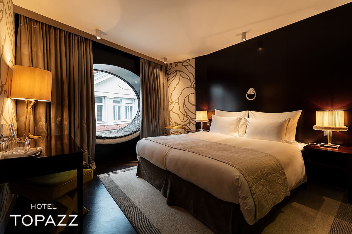 A hotel room with a large bed, two bedside tables with lamps, a view of the outside through a window, and a circular window.