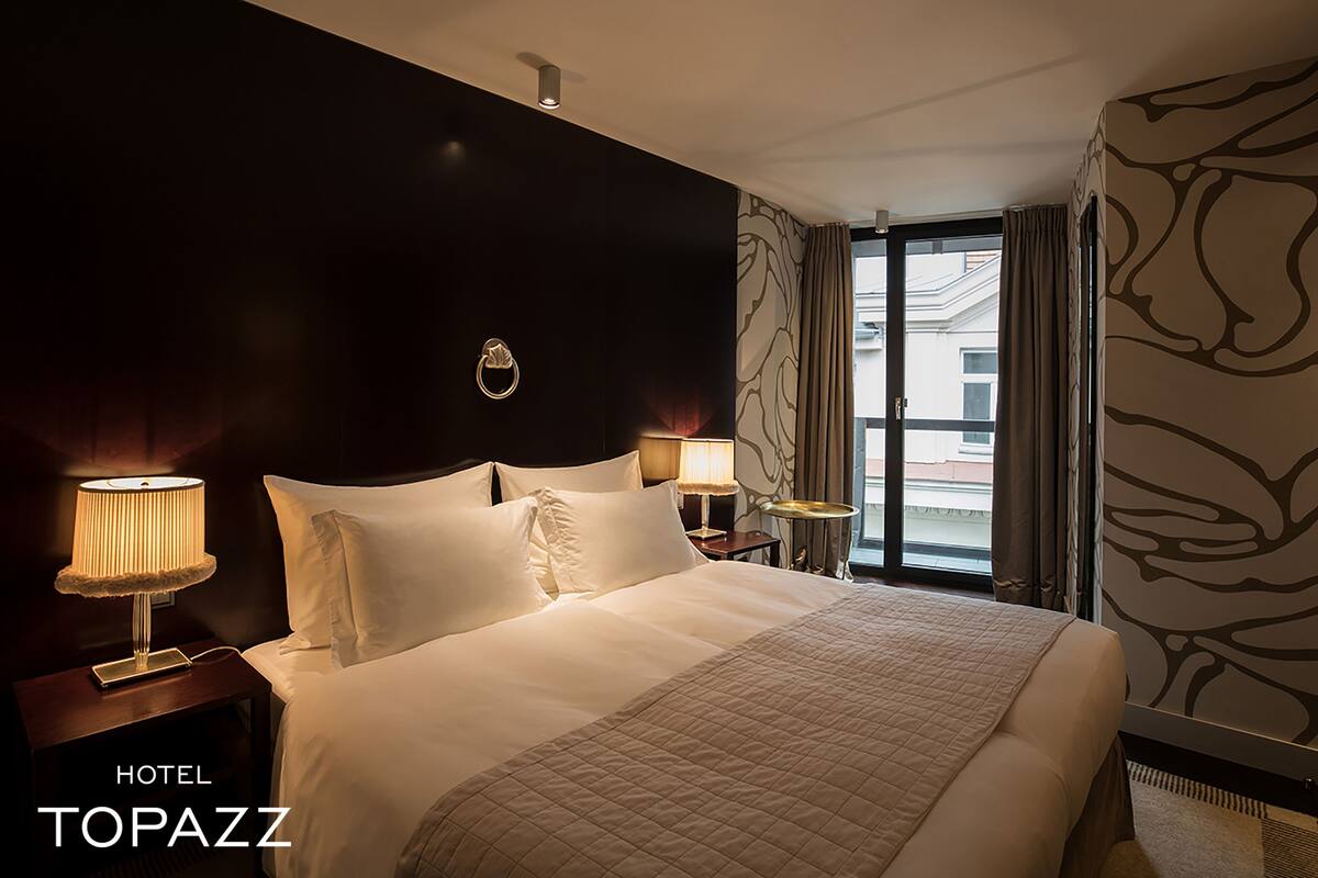 A hotel room with a large bed, two bedside tables with lamps, a headboard with a circular design, and a window with curtains.