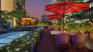 A rooftop bar with outdoor seating, a pool, and city views at dusk.