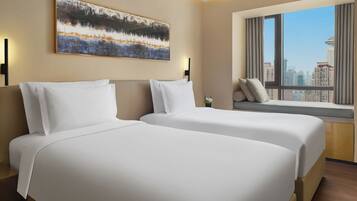 A hotel room with two beds, a large window with a city view, a painting on the wall, and a small plant on the windowsill.