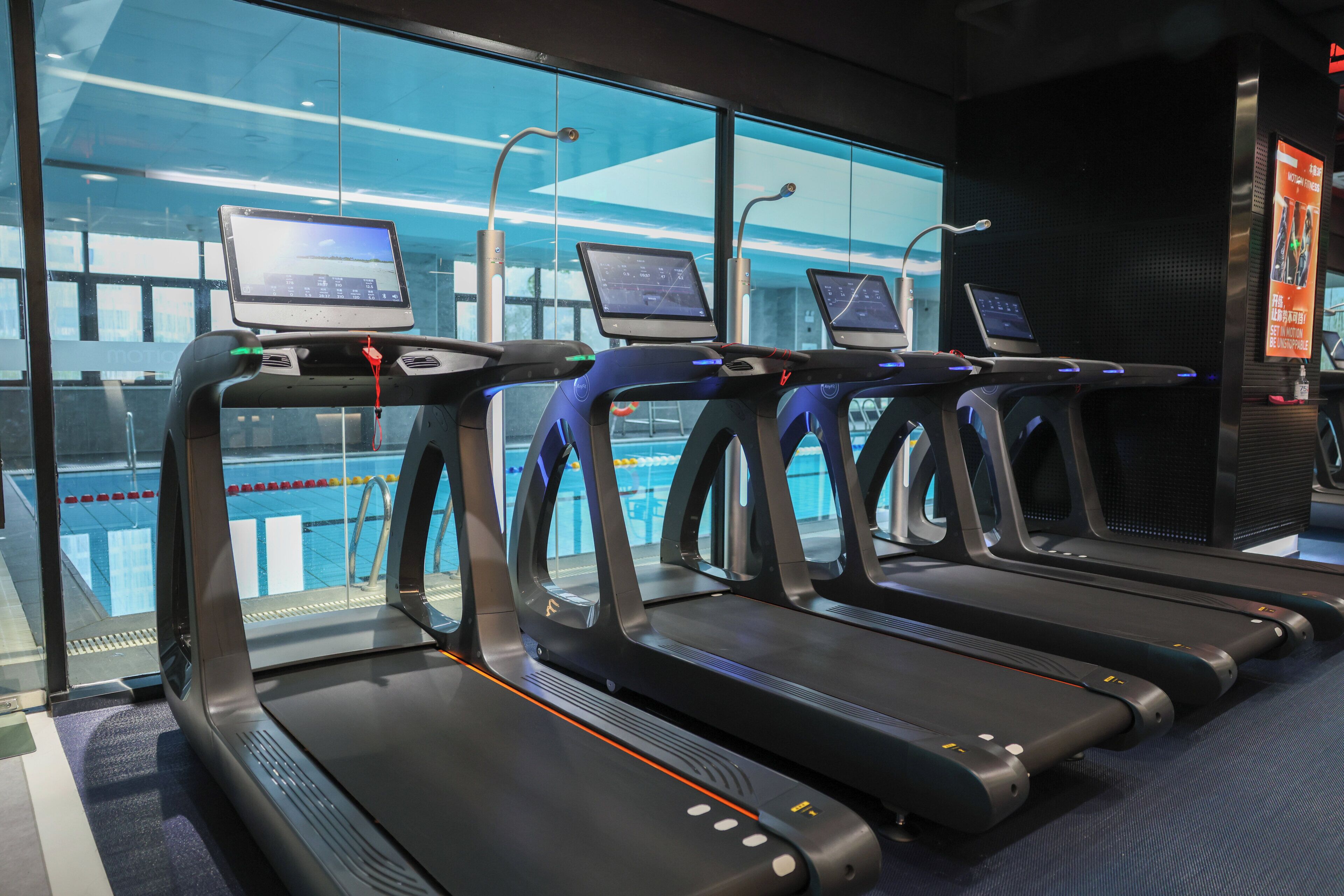 Four treadmills with digital screens, positioned near a window overlooking a pool.