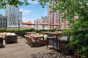 Un patio al aire libre con suelo de madera, mesas, sillas y sombrillas, rodeado de follaje y vistas a un paisaje urbano con edificios altos.