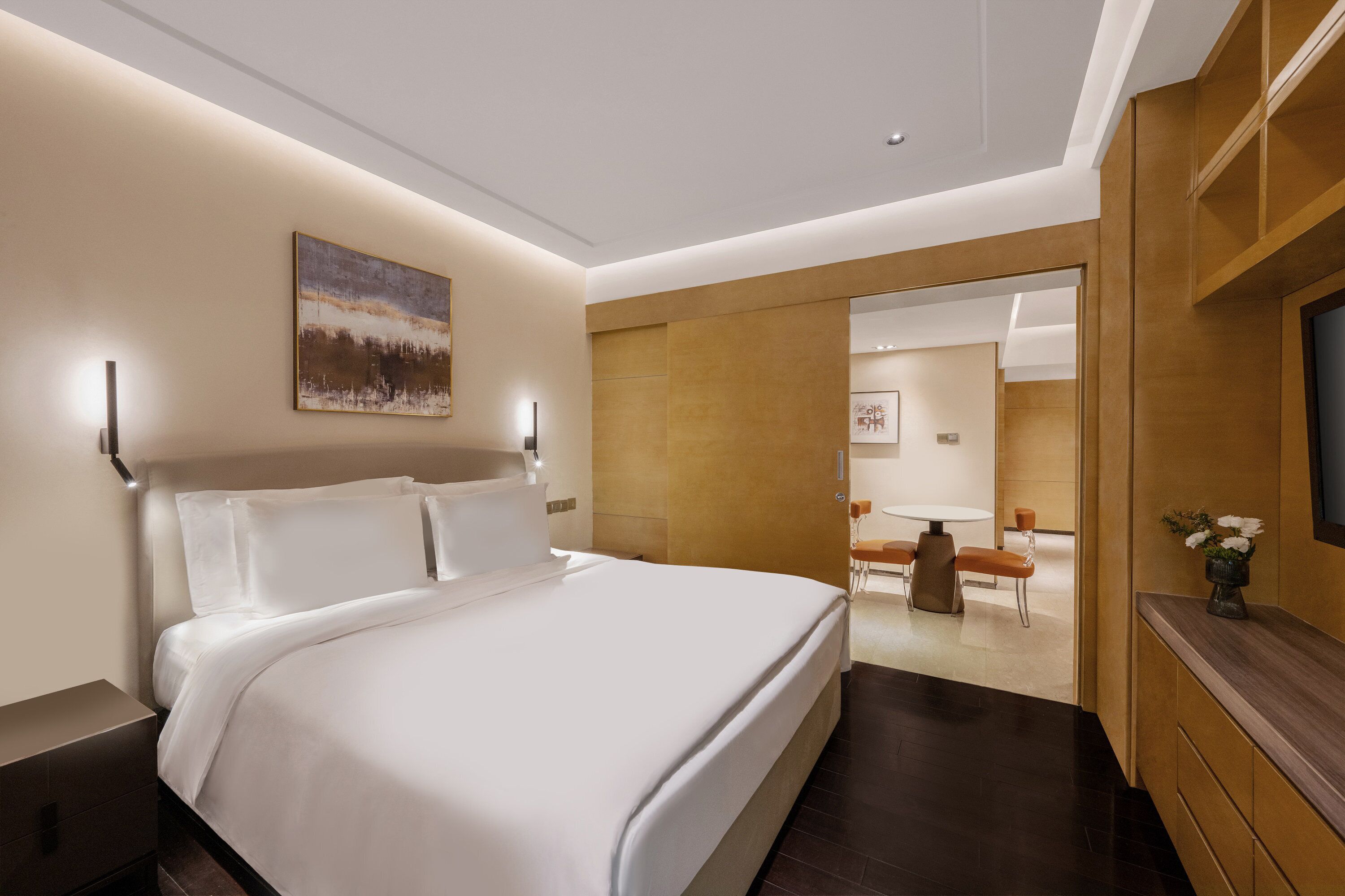 A modern hotel room with a large bed, a painting on the wall, and a view into the dining area.