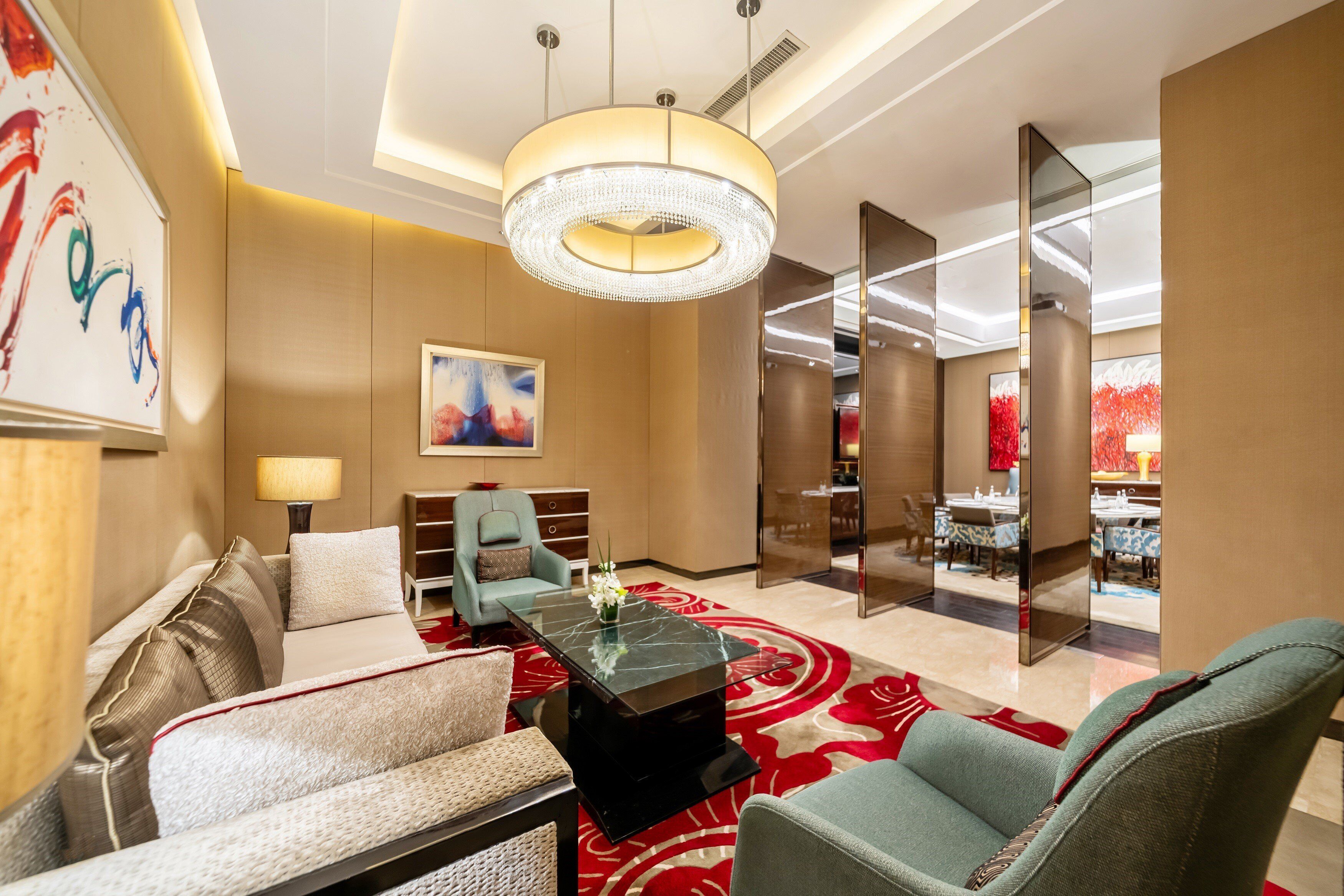 A modern hotel lobby with a marble coffee table, comfortable seating, a large chandelier, and a dining area in the background.