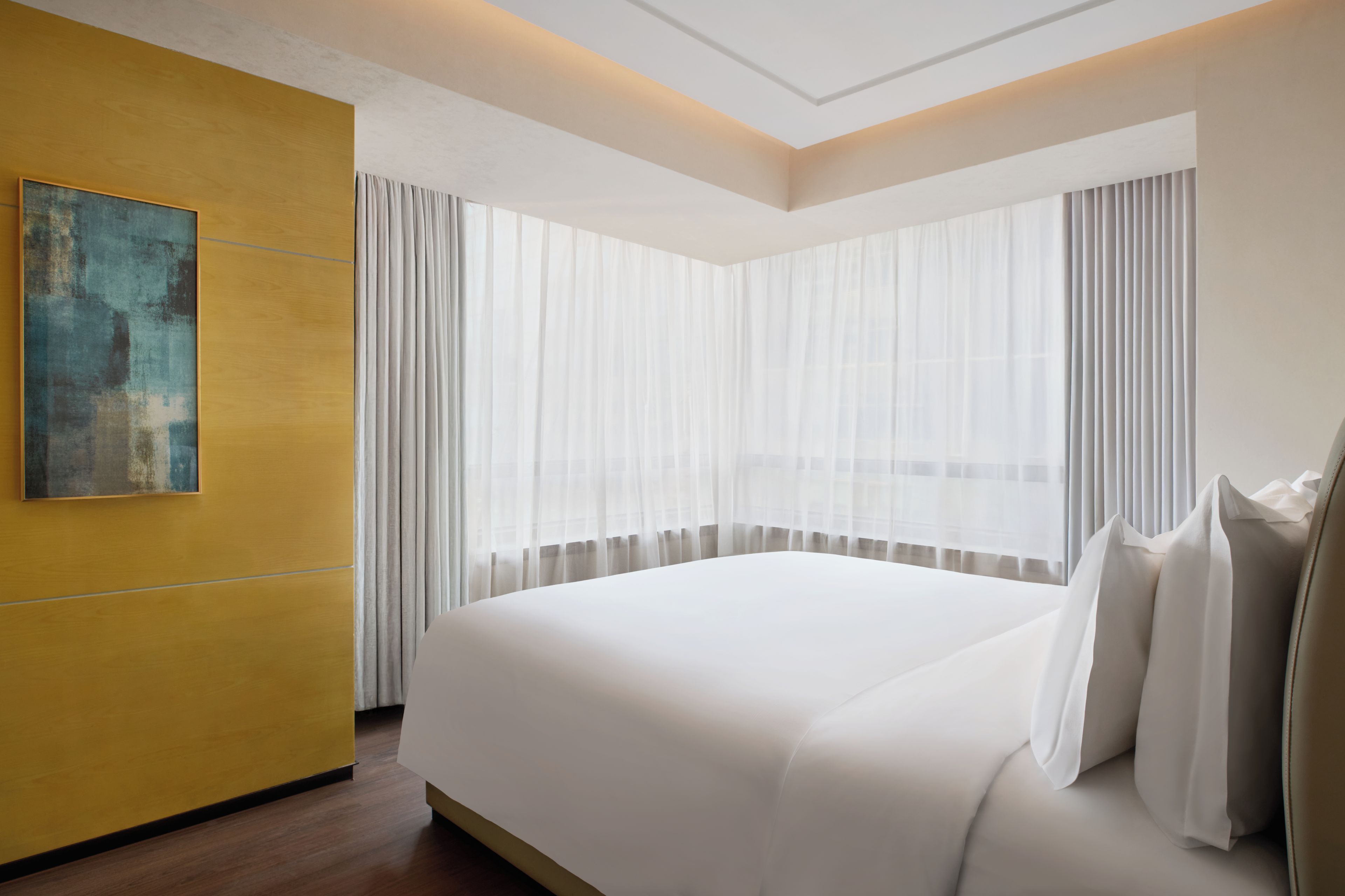 A hotel room with a large bed, a painting on the wall, and sheer curtains.