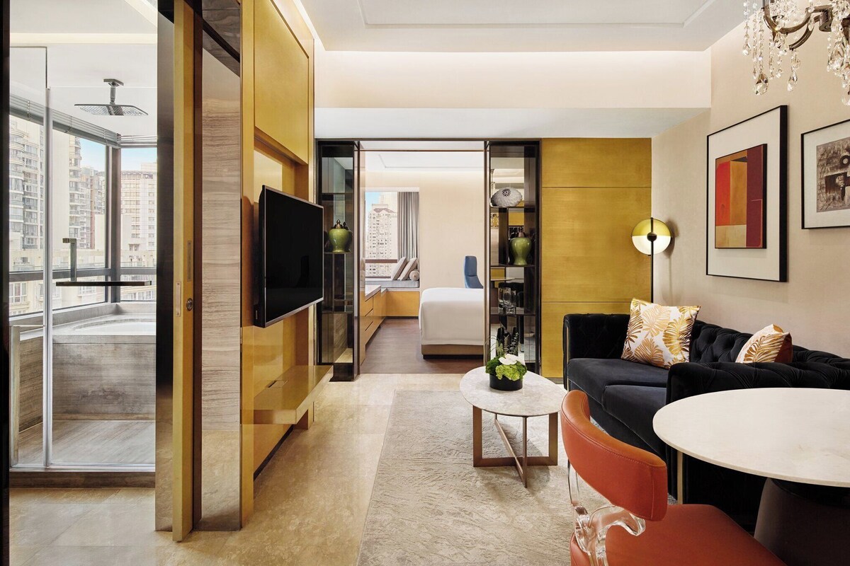 Habitación de hotel moderna con un ventanal grande, televisión de pantalla plana, sofá, mesa de centro y zona de comedor.