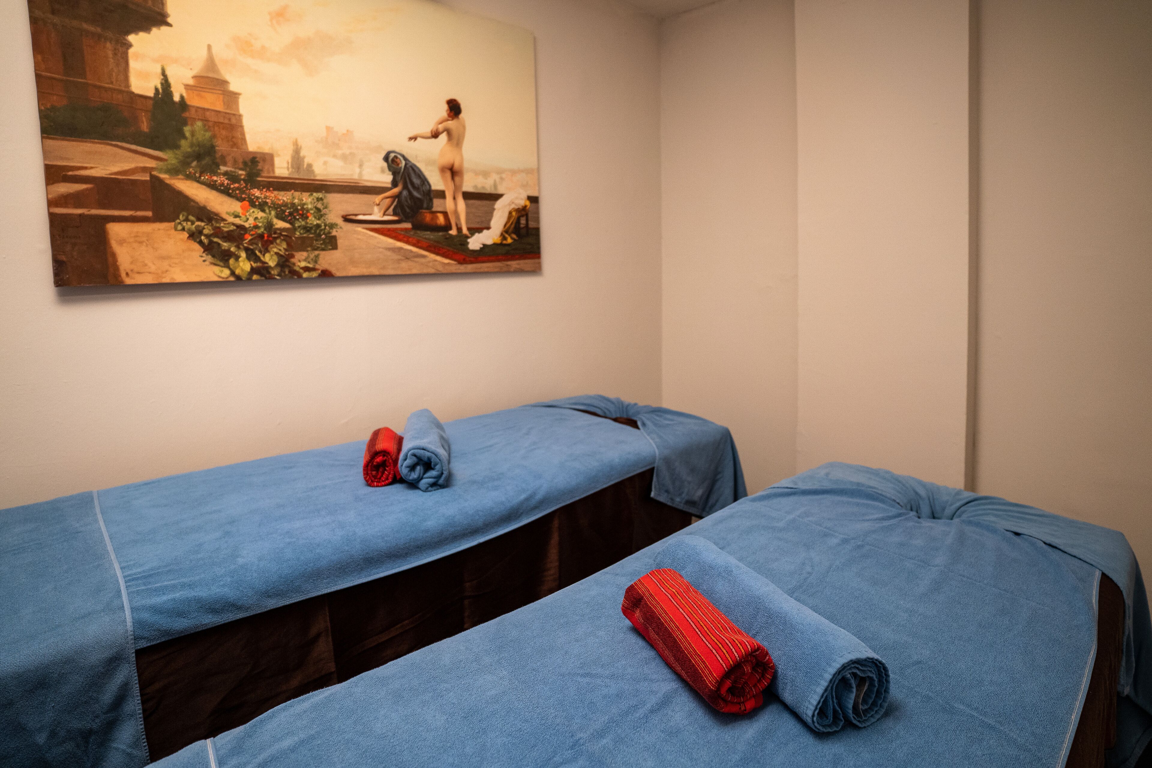 A room with two massage tables covered in blue sheets, one with a red towel, and a large painting on the wall depicting a historical scene.
