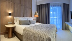 A modern hotel room with a large bed, a bedside table, a wall-mounted TV, and a view of a building through a window with curtains. - Bodrum Beach Resort (Bodrum)