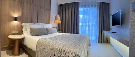 A modern hotel room with a large bed, a bedside table, a wall-mounted TV, and a view of a building through a window with curtains.