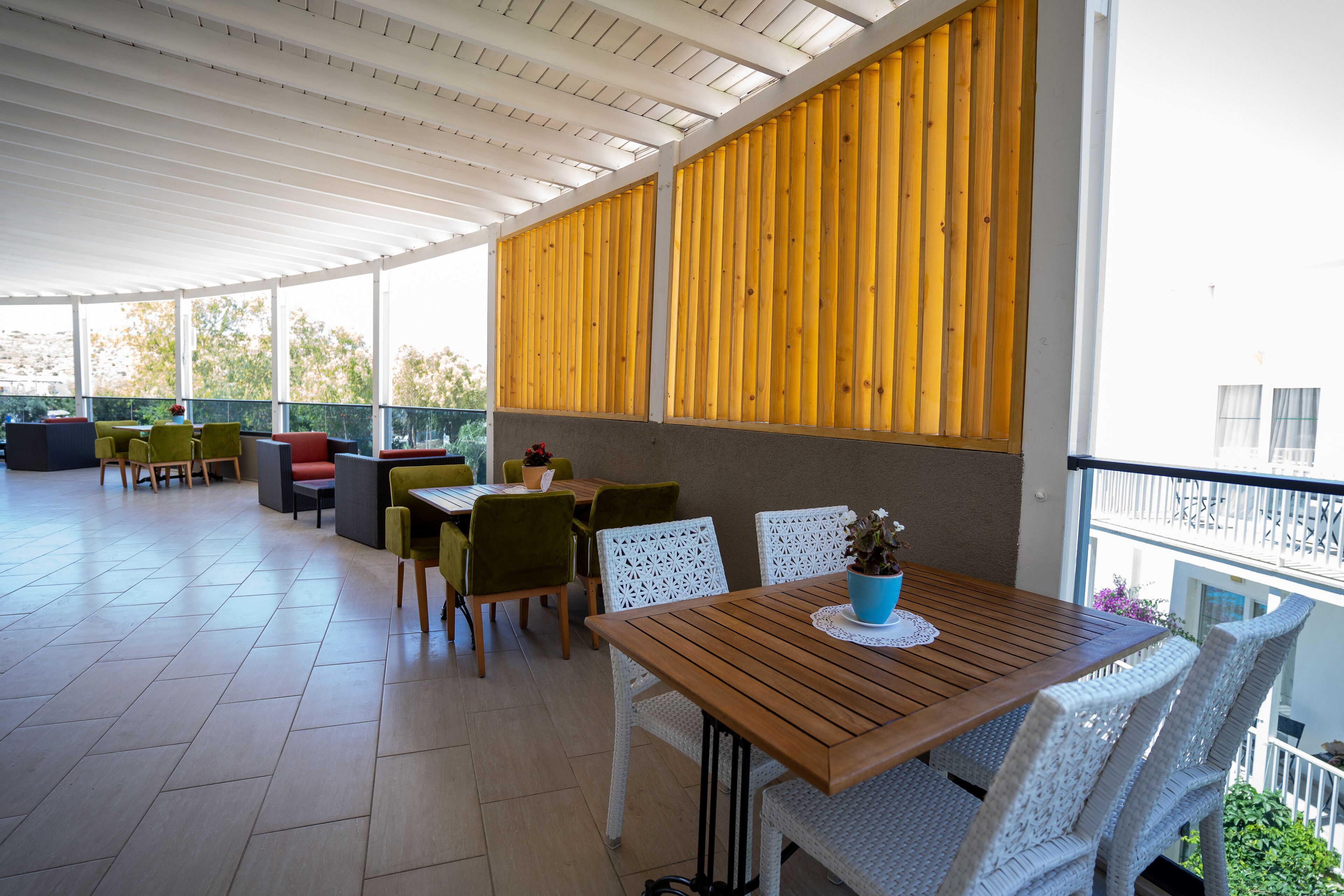 A covered outdoor dining area with a wooden table and white wicker chairs, surrounded by a white railing and large windows.