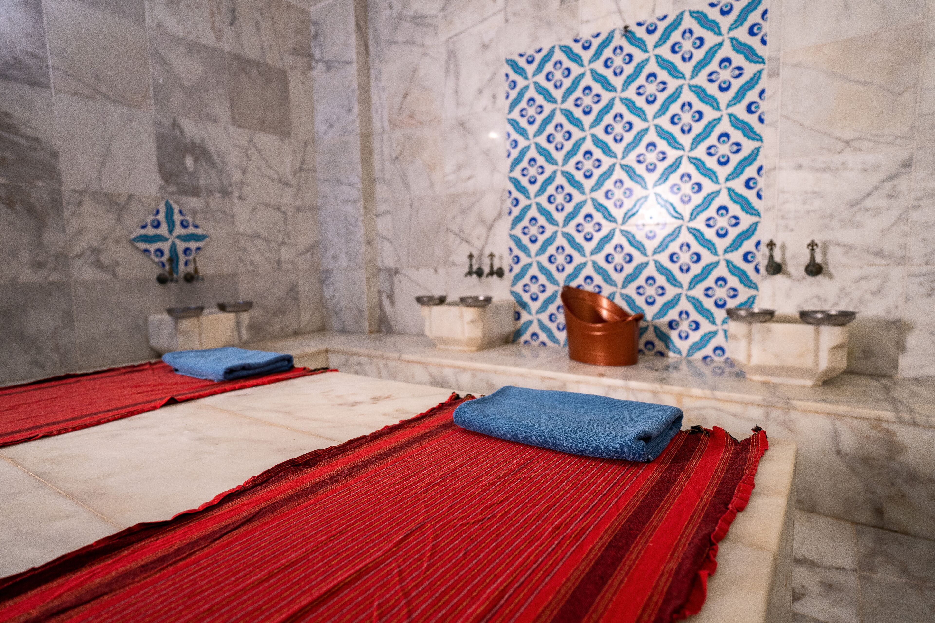 Une salle de bain aux murs recouverts de marbre, avec un rideau de douche aux motifs rouges et bleus, et deux vasques dotées de robinets en cuivre.