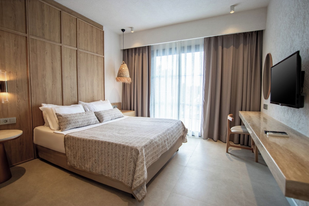 A modern hotel room with a large bed, a wooden panel wall, a desk with a chair, a TV, and a large window with curtains.