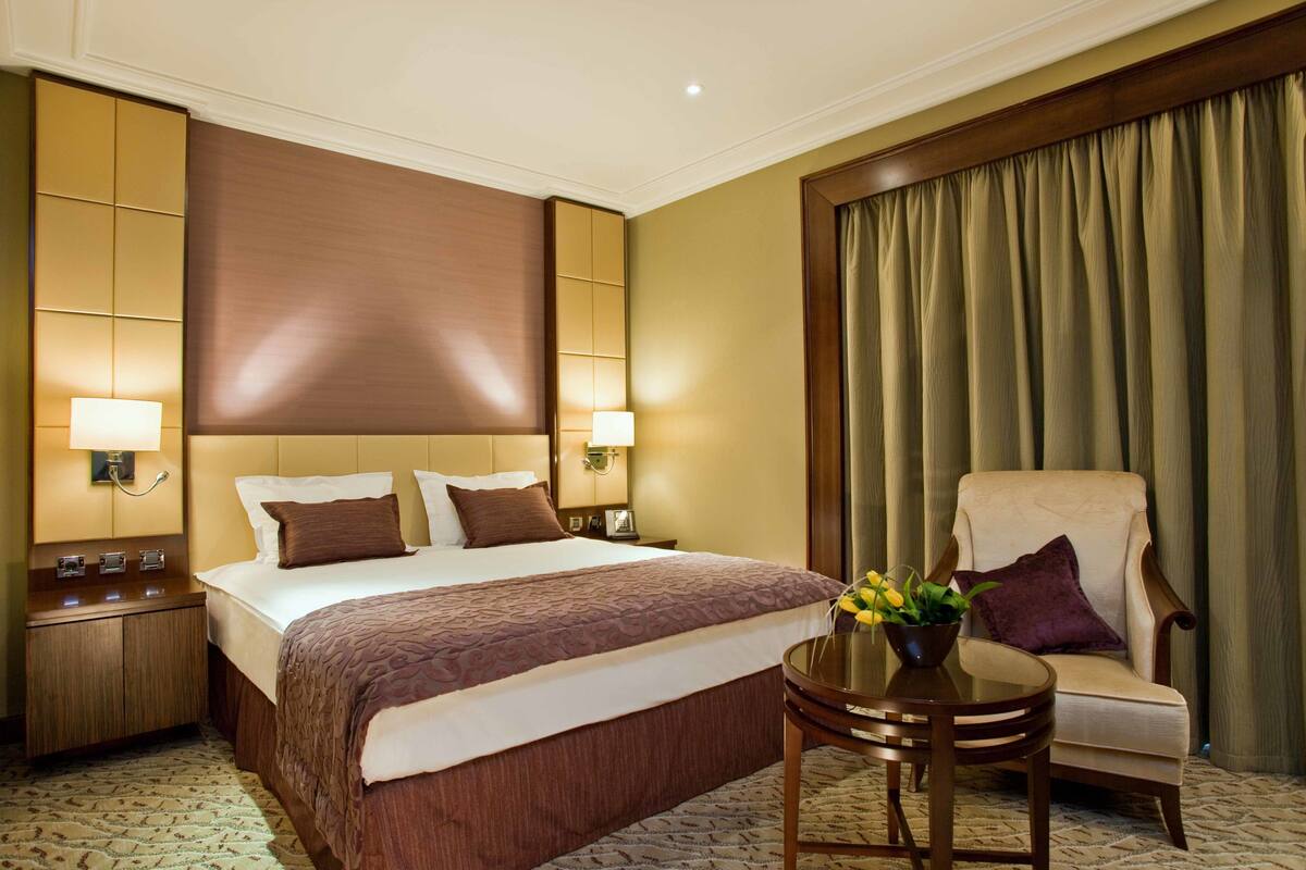 A hotel room with a large bed, two bedside tables with lamps, a round table with a vase of flowers, a chair, and a large window with curtains.