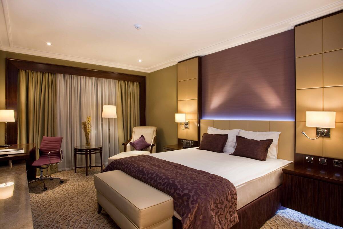 A hotel room with a large bed, two bedside tables, a chair, a desk, and a small table with a vase.