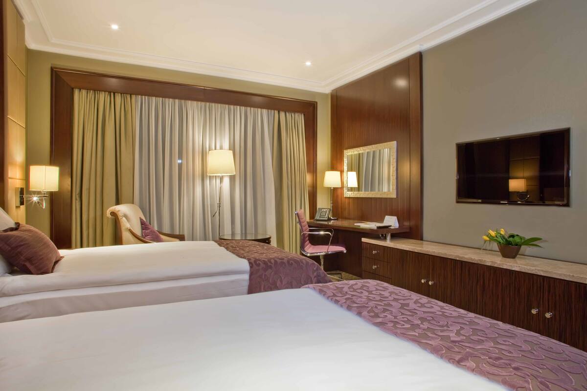 A hotel room with two beds, a desk, a chair, a TV, and a large window with curtains.