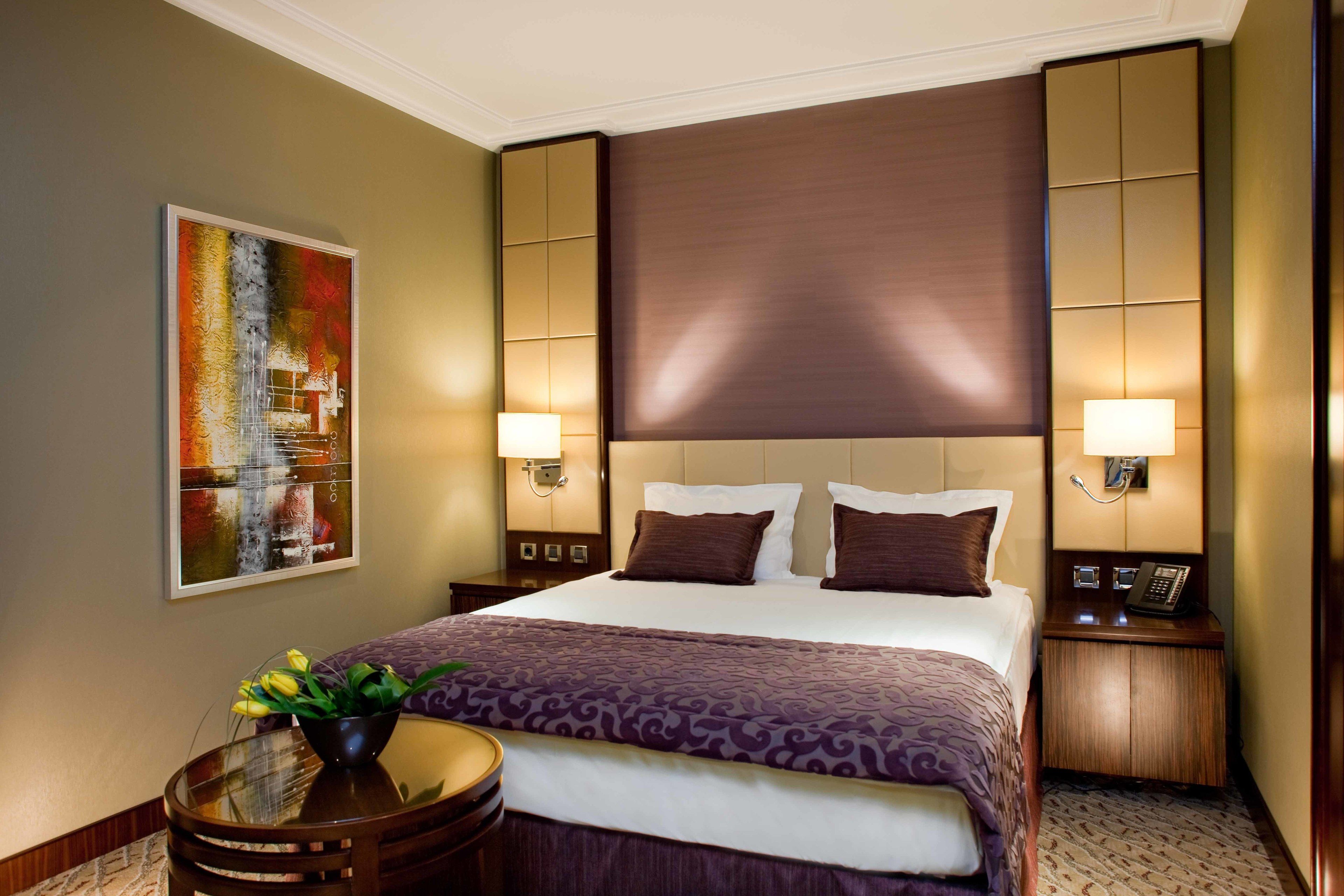 Una habitación de hotel con una cama grande, dos mesitas de noche con lámparas, una obra de arte en la pared y una pequeña mesa redonda con un florero.