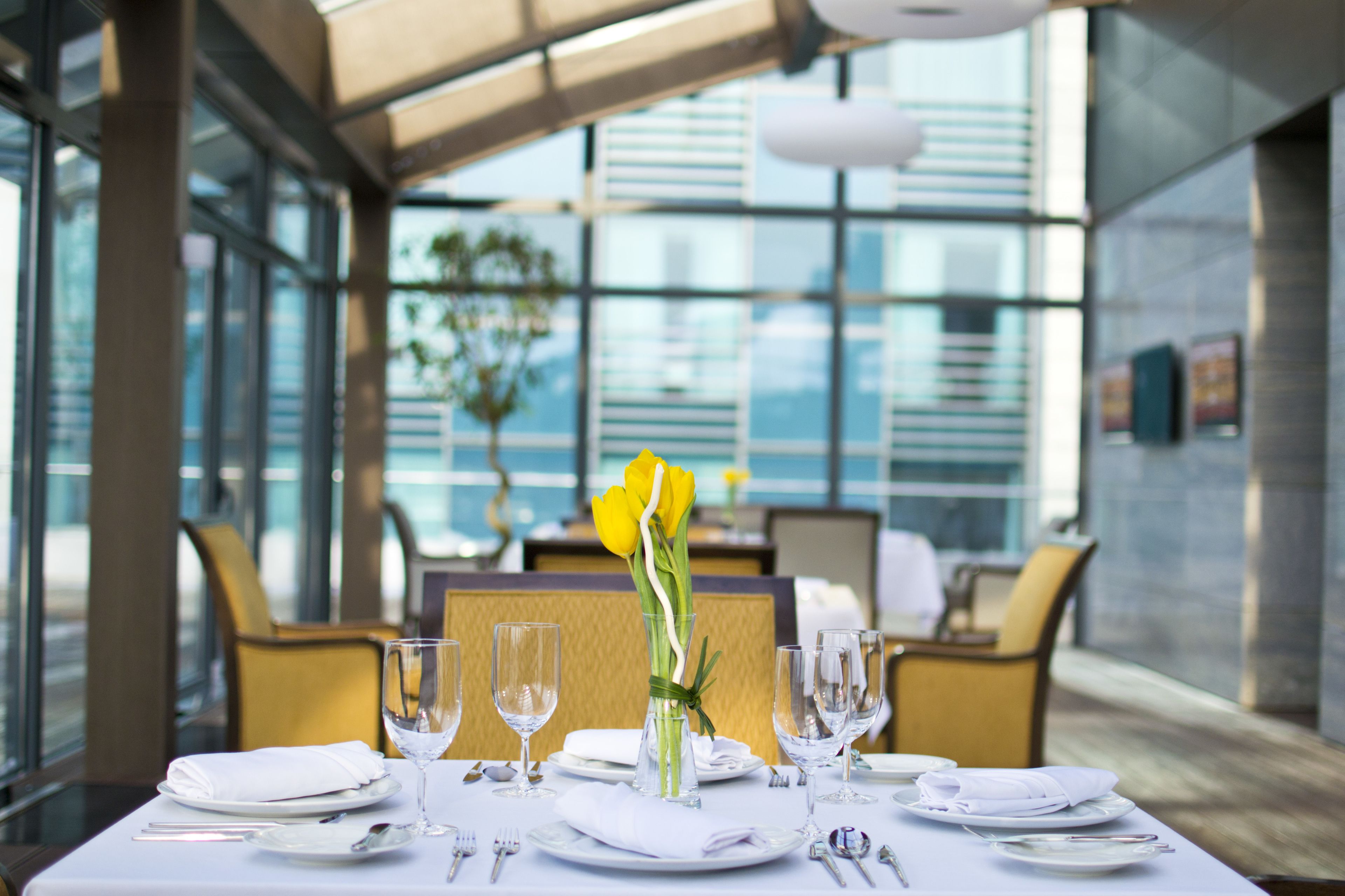 A dining table set for a meal with a vase of yellow flowers, clear glasses, and cutlery.