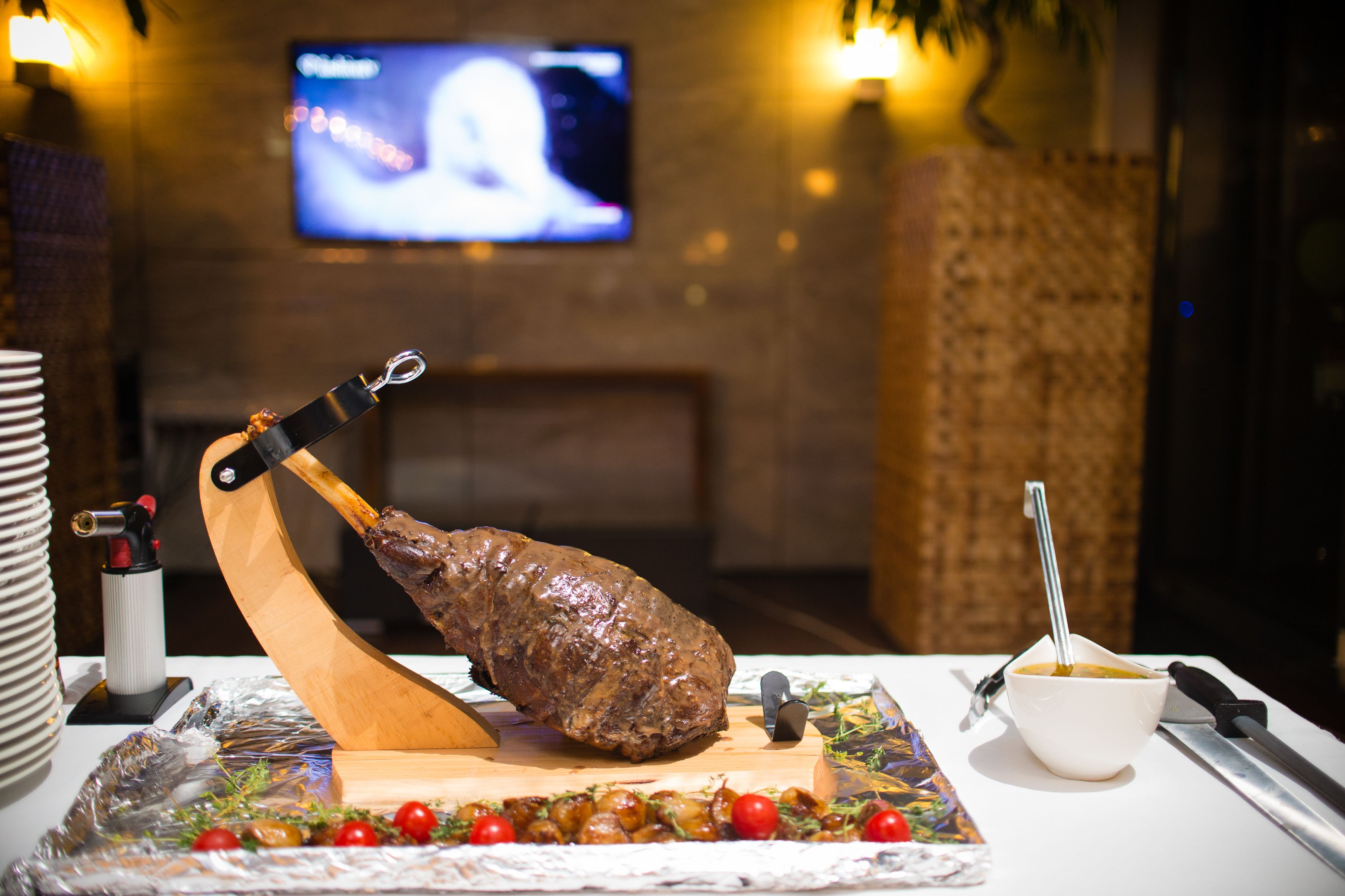 A large roasted meat centerpiece on a wooden board, a bowl of soup, and a knife on a table with a decorative mat.