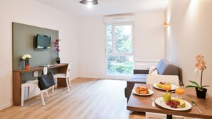 A modern room with a wooden desk, a television, a dining table with breakfast, a sofa, and a large window.