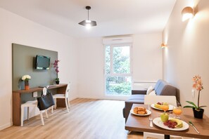 A modern room with a wooden desk, a television, a dining table with breakfast, a sofa, and a large window. - Comfort Aparthotel Versailles Saint Cyr l'Ecole (Saint-Cyr-l'Ecole)