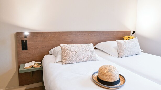 A neatly made bed with two pillows, a bedside table with a book and a phone, and a straw hat on the bed.