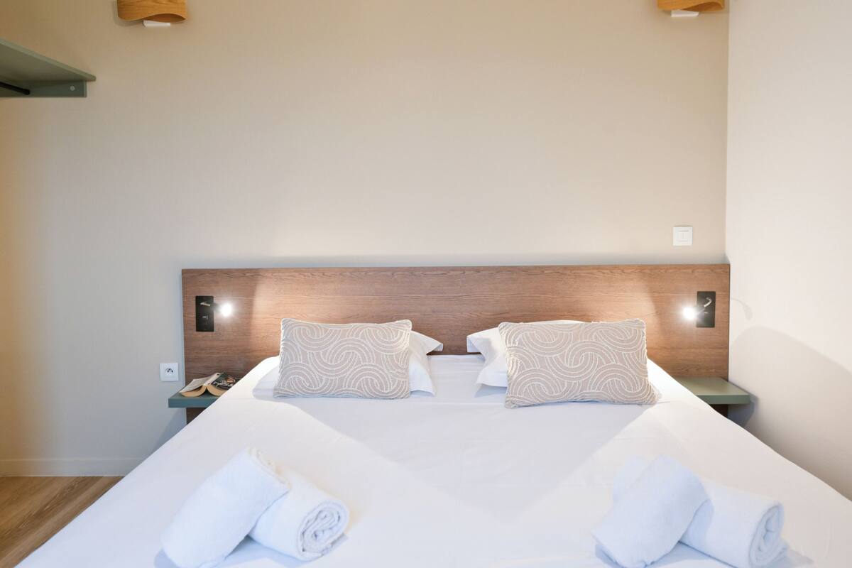 A neatly made bed with two pillows, a wooden headboard, and a bedside table with a phone and a small item on it.