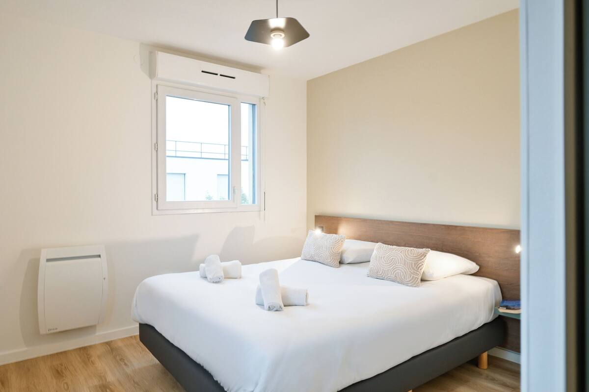 A modern hotel room with a large bed, a window, and a bedside table.
