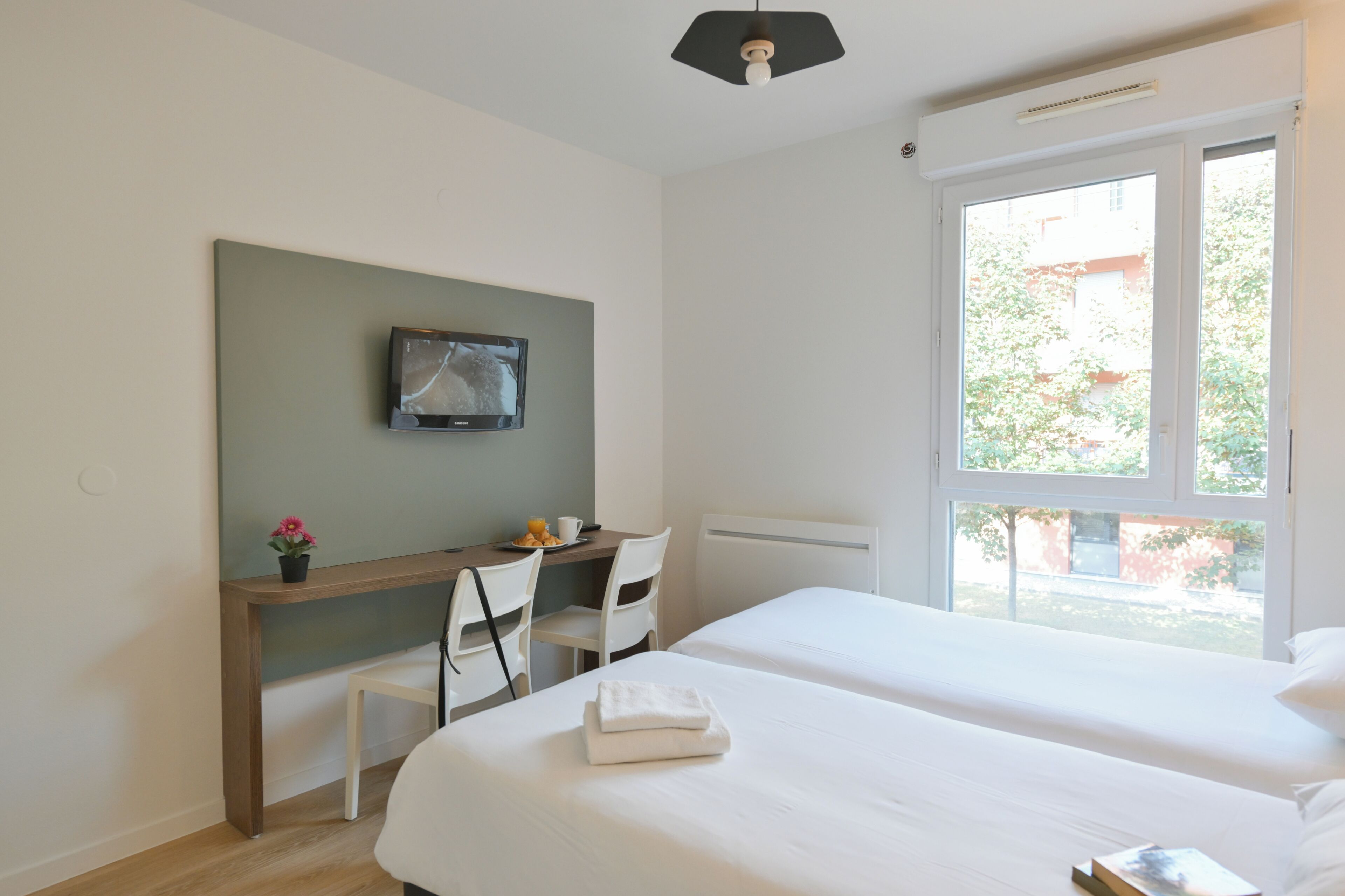 A hotel room with two beds, a desk with a chair, a TV mounted on the wall, and a window with a view of greenery outside.