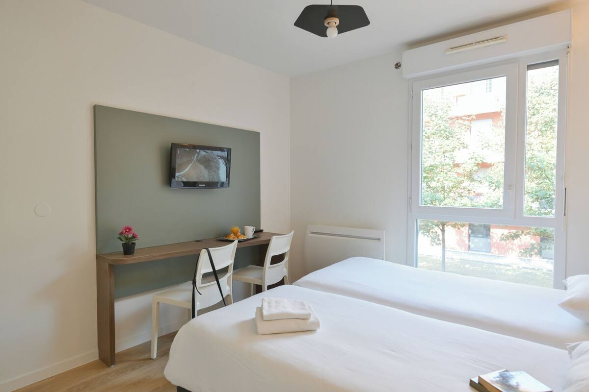 A hotel room with two beds, a desk with a chair, a TV mounted on the wall, and a window with a view of greenery outside.