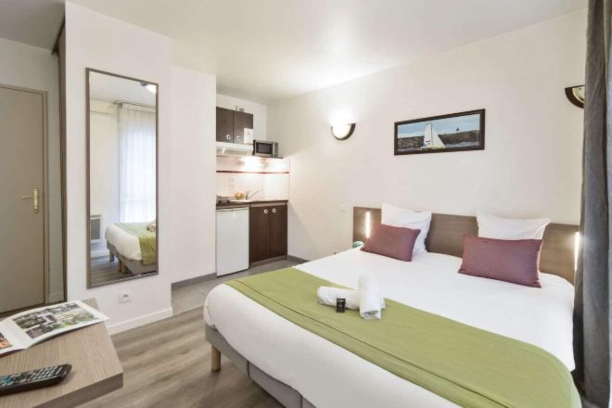 A hotel room with a bed, a desk, a mirror, a picture on the wall, and a kitchenette.