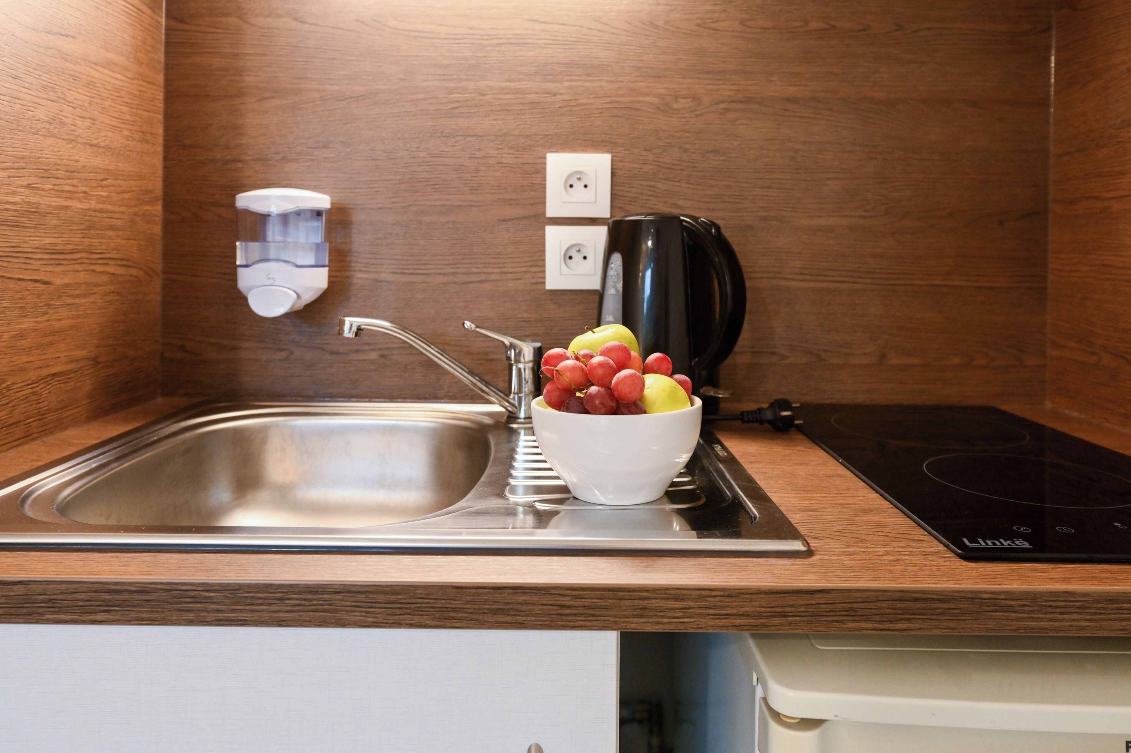 A modern kitchen with a stainless steel sink, a bowl of fruit, a kettle, and a Linksys Wi-Fi router.