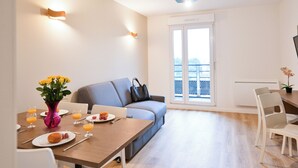 A modern living room with a sofa, a dining table with breakfast items, and a balcony with a view.